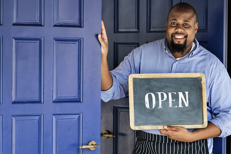 Business Insurance - Restaurant Business Owner Standing by Blue Doors and Wearing a Smok is Smiling and Holding an Open Sign
