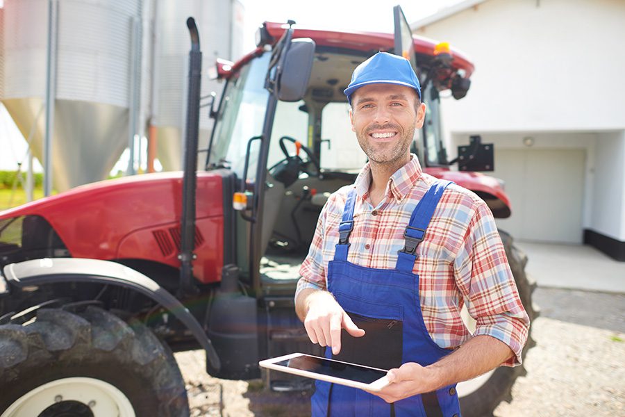 Specialized Business Insurance - A Farmer Wearing Blue Overalls is Smiling and Holding a Tablet While Standing in Front of His Red Tractor at His Farm on a Sunny Day